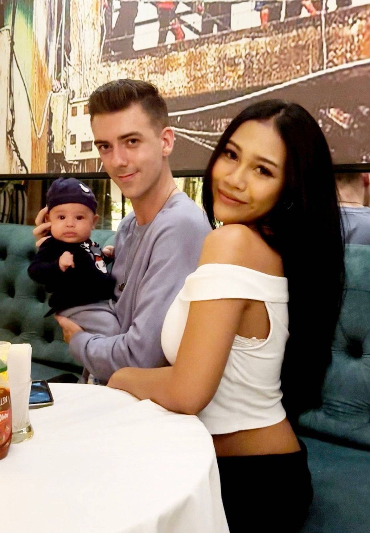 បុរស​និង​ស្ត្រី​ពេញវ័យ​កំពុង​អង្គុយ​ជាមួយ​គ្នា​នៅលើ​កៅអី។ បុរស​កំពុង​តែ​កាន់​កូន ហើយ​ស្ត្រី​កំពុង​ញញឹម។ ពួកវាត្រូវបានហ៊ុំព័ទ្ធដោយតុ និងជញ្ជាំងដែលមានផ្ទាំងគំនូរ។